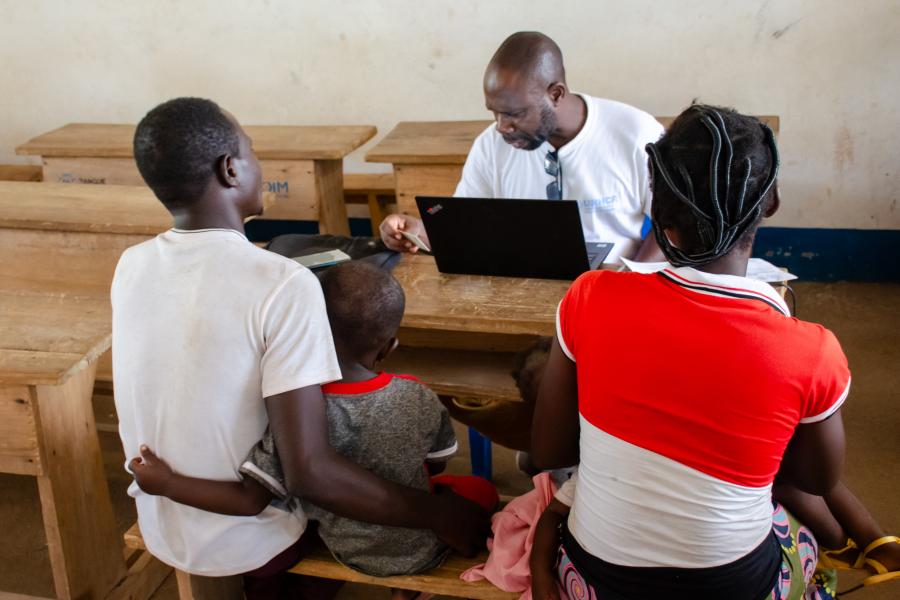 A family group sit at a desk with a UNHCR staff member and a laptop