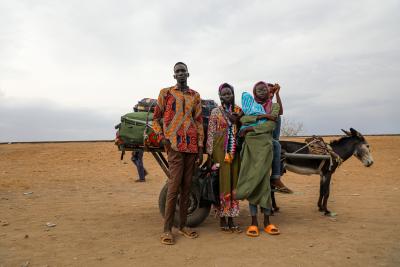 A small group of people, one holding a young child, stand next to a loaded cart and donkey.