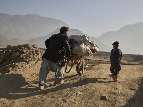 A man is pushing a wheelbarrow full of firewood up a hill. He is accompanied by his five-year-old son walking next to him.