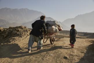 A man is pushing a wheelbarrow full of firewood up a hill. He is accompanied by his five-year-old son walking next to him.