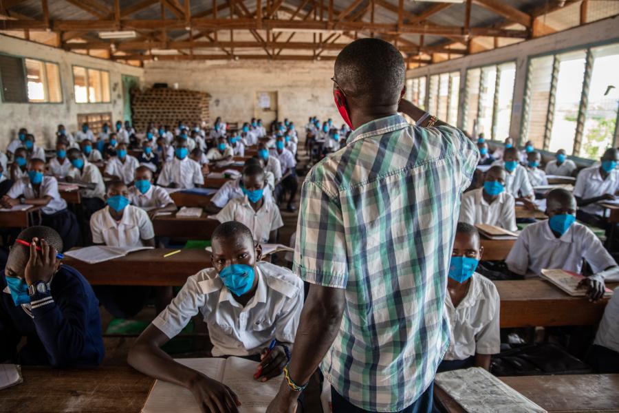 Rear view of a professor actively teaching a full class of learners. The students facing the camera are wearing blue COVID-19 masks and school uniforms.