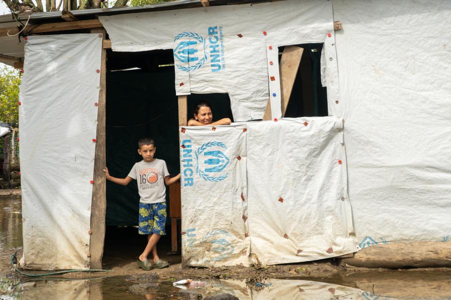 A boy standing on the entrace of their makeshift house while with his mother watching through a window