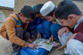 Khalil, in a white hat, a third grade student in an Afghan refugee school in Pakistan reading a story from his book to Mehmood (in Black Dress) who is out of school.