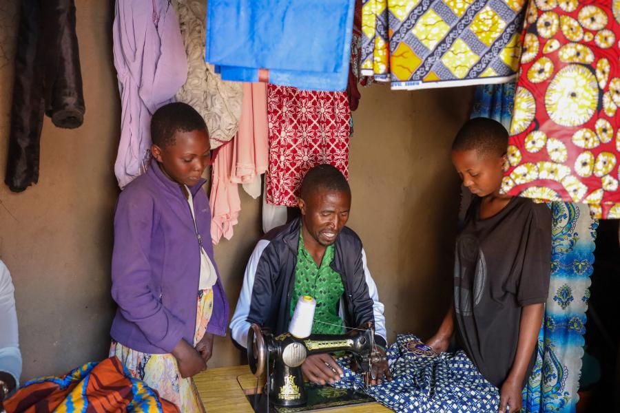 A group of three people with clothes hanging around them. The middle person sits at a table and works on a sewing machine while the other two people who are younger, watch his instruction.